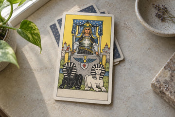 A printed Chariot tarot card with an armored driver and two sphinxes on a sunlit stone desk.